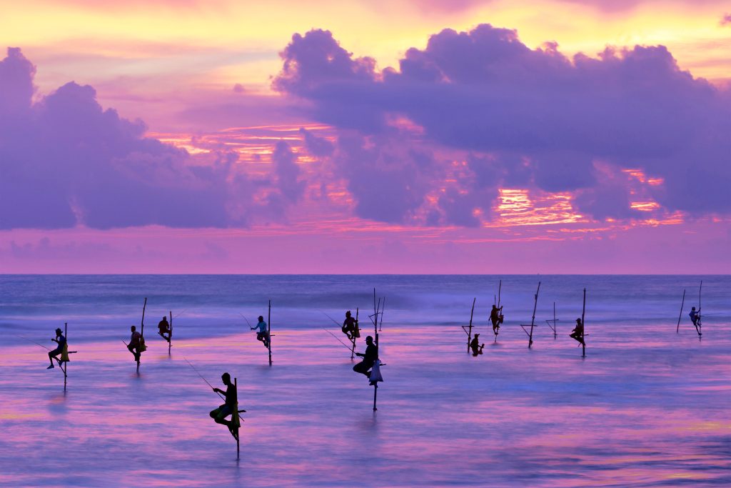 A sunset photo showing silhouetted figures balancing on stilts in the water holding fishing lines